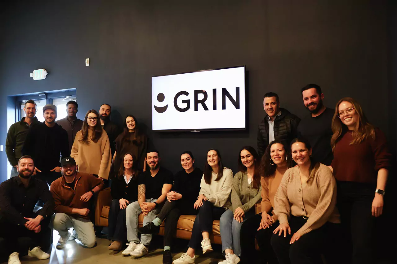 A group of people, embodying the essence of Grin Careers, are seated and standing in front of a dark wall showcasing the word GRIN. Casually dressed, some are lounging on a couch while others stand behind them, all sharing cheerful smiles with the camera.
